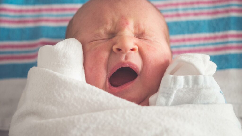 Neugeborenes Baby in weißer Decke gähnt mit geschlossen Augen, im Hintergrund blau-weiß gestreifter Stoff.