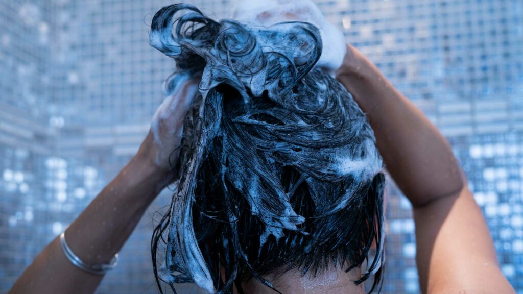 Eine Frau shampooniert sich die Haare in der Dusche. In ihren Haaren ist viel weißer Schaum.