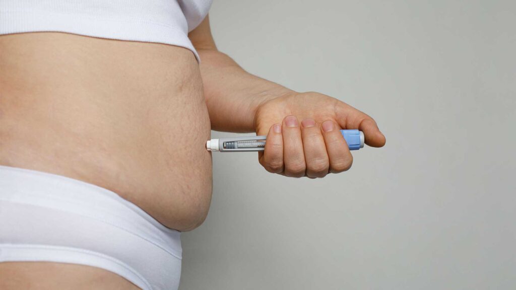 Mensch mit weißer Unterwäsche spritzt sich eine Spritze in den nackten Bauch