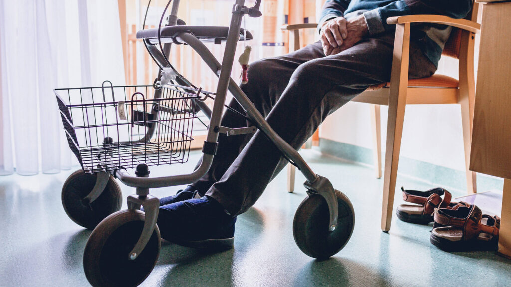 Ein älterer Mensch sitzt mit gefalteten Händen auf einem Stuhl, ein Rollator mit Korb steht davor. Im Hintergrund sind Hausschuhe und Möbel in einem lichtdurchfluteten Raum sichtbar.