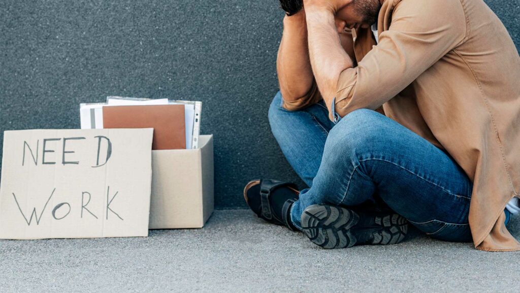 Ein Mann sitzt verzweifelt hinter einem Schild aus Pappe mit der Aufschrift "Need Work".