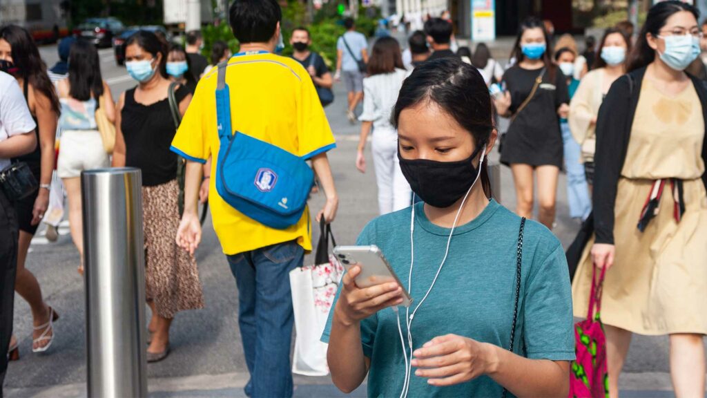Fußgänger mit Mundschutz gehen die Orchard Road im Stadtzentrum von Singapur entlang. Alle Menschen tragen Masken, im Vordergrund ist eine Frau zu sehen, die auf ihr Handy schaut.
