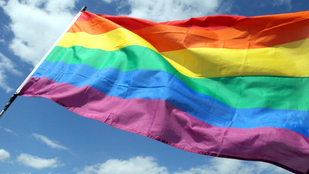 Wir sehen die vor einem blauen Himmel wehende Regenbogenflagge.