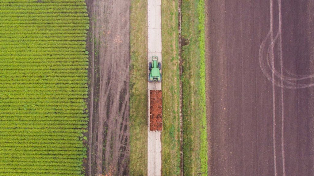Ein Traktor mit Anhänger fährt zwischen zwei Äckern auf einer Straße.