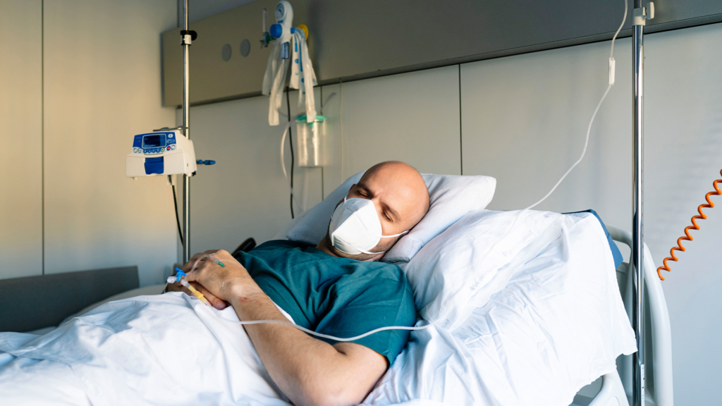 Ein Mann liegt mit FFP2 Maske und Tropf in einem Krankenhausbett