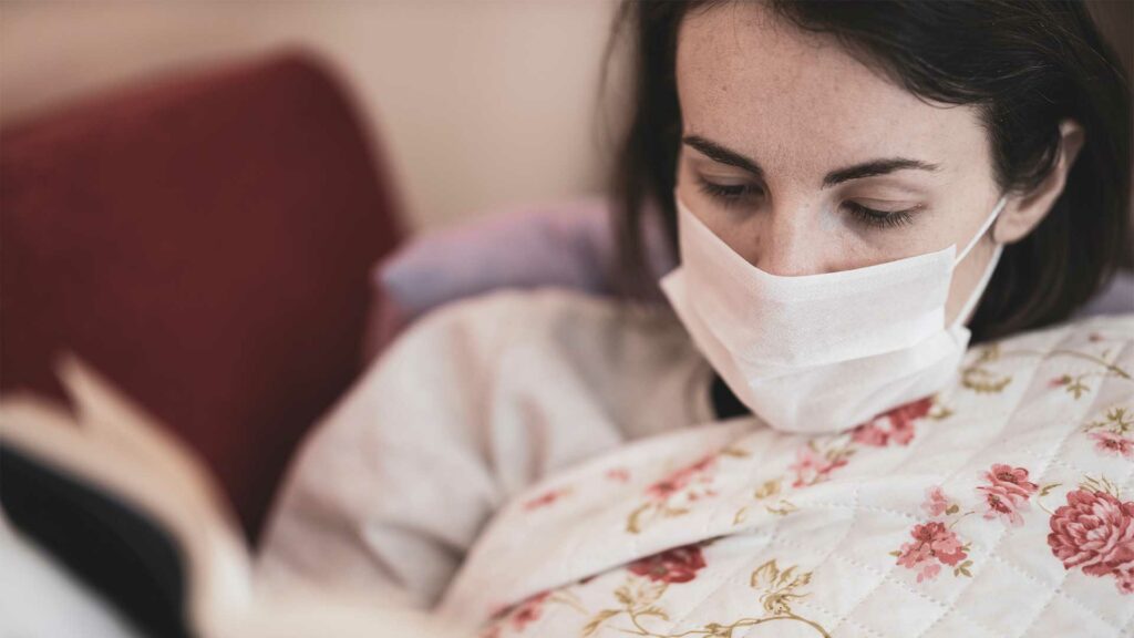 Eine junge Frau liegt im Bett und hat eine Maske auf