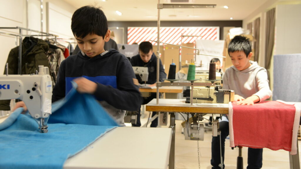 Drei Kinder sitzen hintereinander an Nähmaschinen und arbeiten. Sie wirken konzentriert und tragen Sweatshirts.