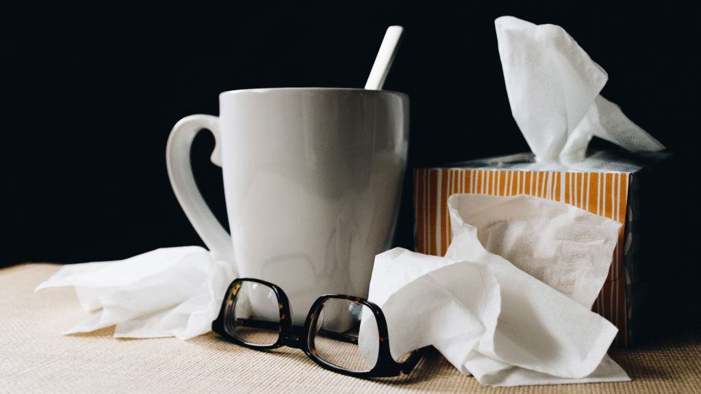 Auf einem Tisch stehen eine Tasse Tee mit einem Löffel, eine abgelegte Brille und eine Packung Taschentücher.