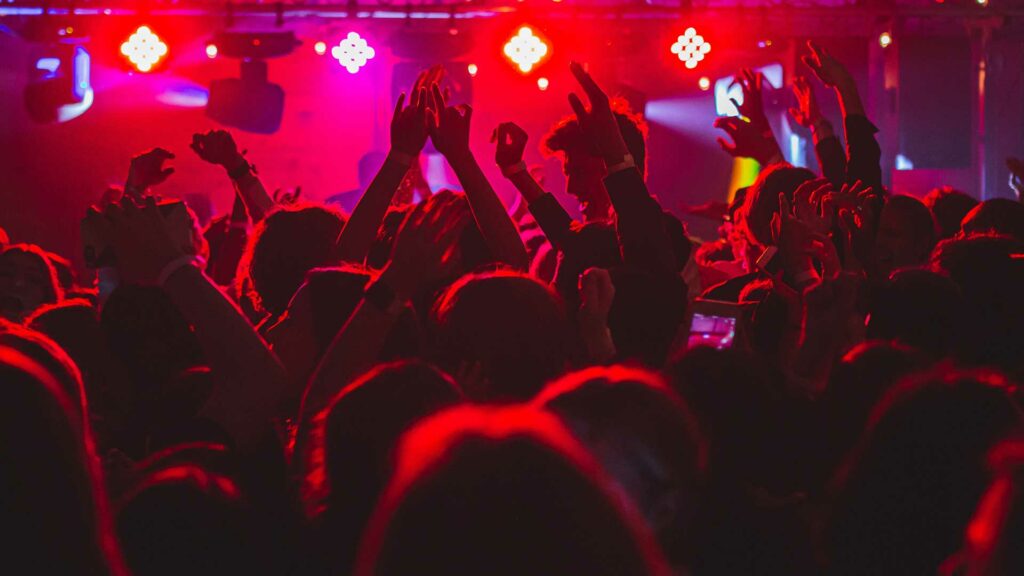 Eine Menschenmenge in einem Club, die Hände jubelnd in die Luft streckt, beleuchtet von roten und violetten Lichtern.