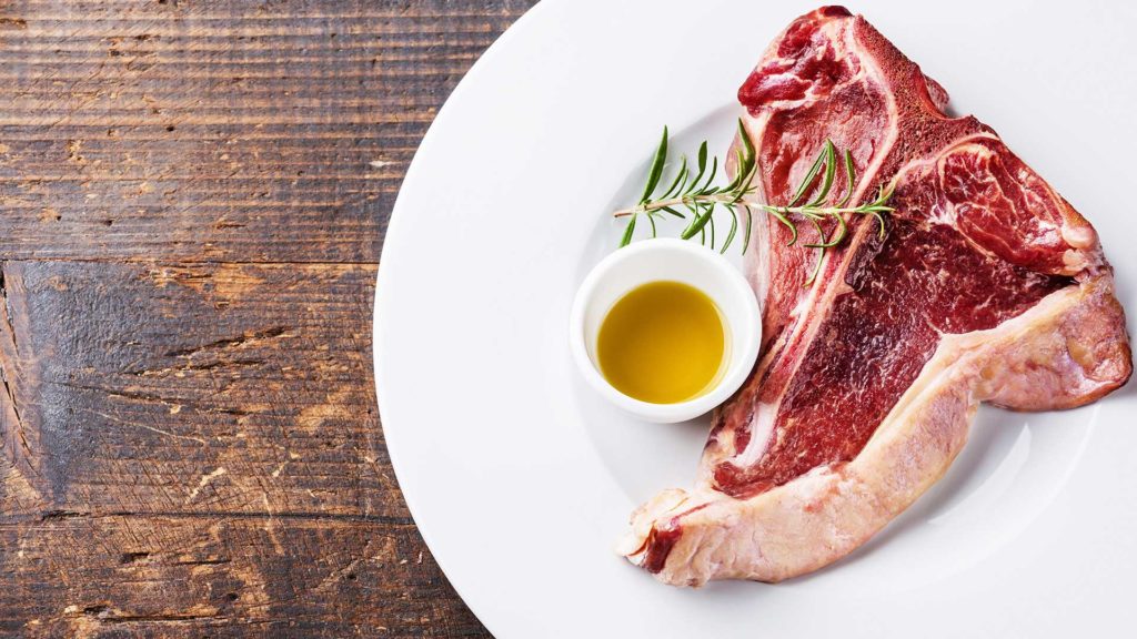 Auf einem weißen Teller liegt ein Stück Fleisch mit einem kleinen Schälchen Öl und einem Rosmarinzweig. Der Teller steht auf einem Holztisch.