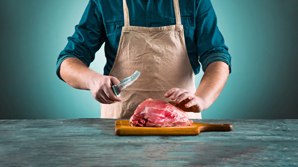 Mann in Grillschürze und Messer in der Hand steht vor einem hölzernen Brett mit rohem Fleisch. Er ist bereit das Fleisch zu verarbeiten.