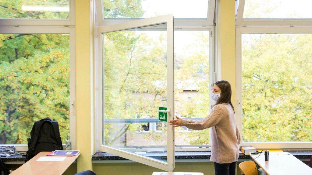 Ein Mädchen mit Maske öffnet im Klassenzimmer das Fenster.