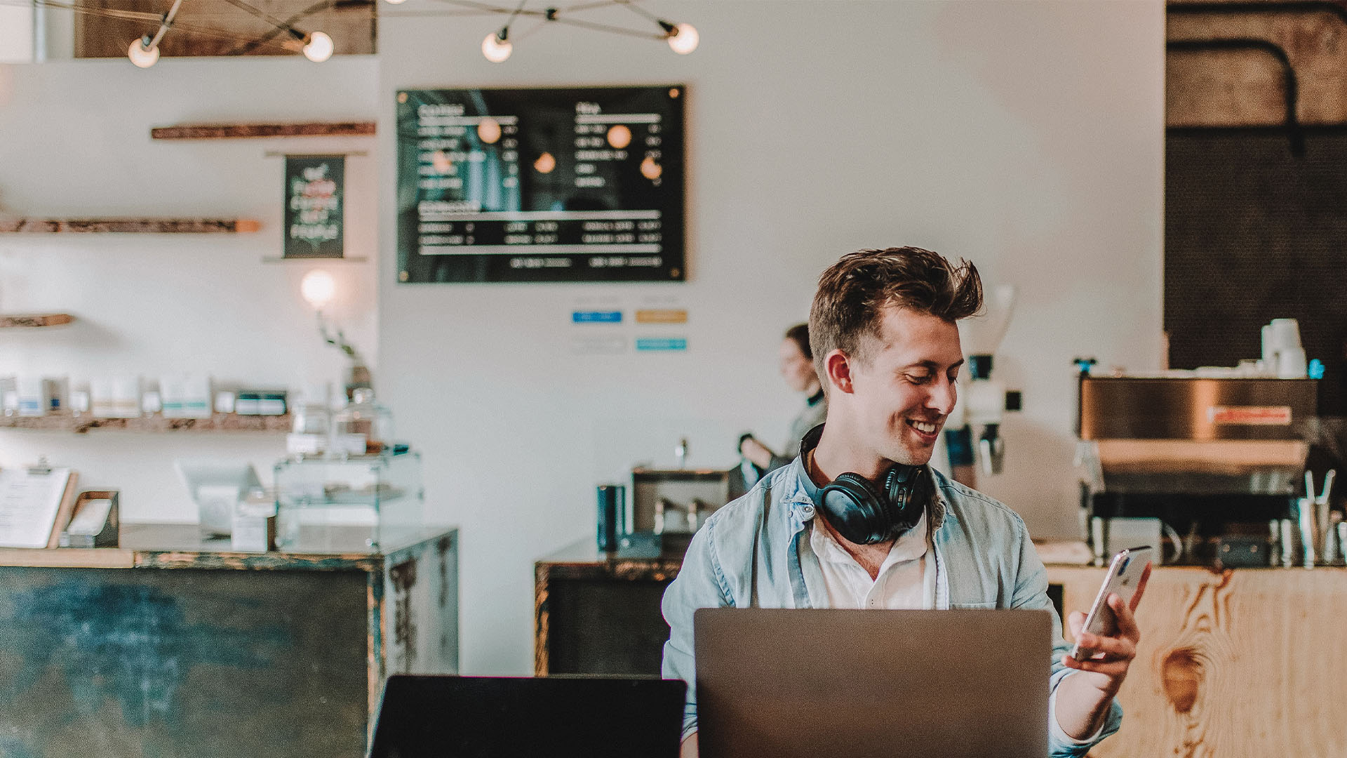 Ein junger Mann sitzt mit Laptop und Smartphone in einem Cafe und schaut lächelnd auf sein Telefon.