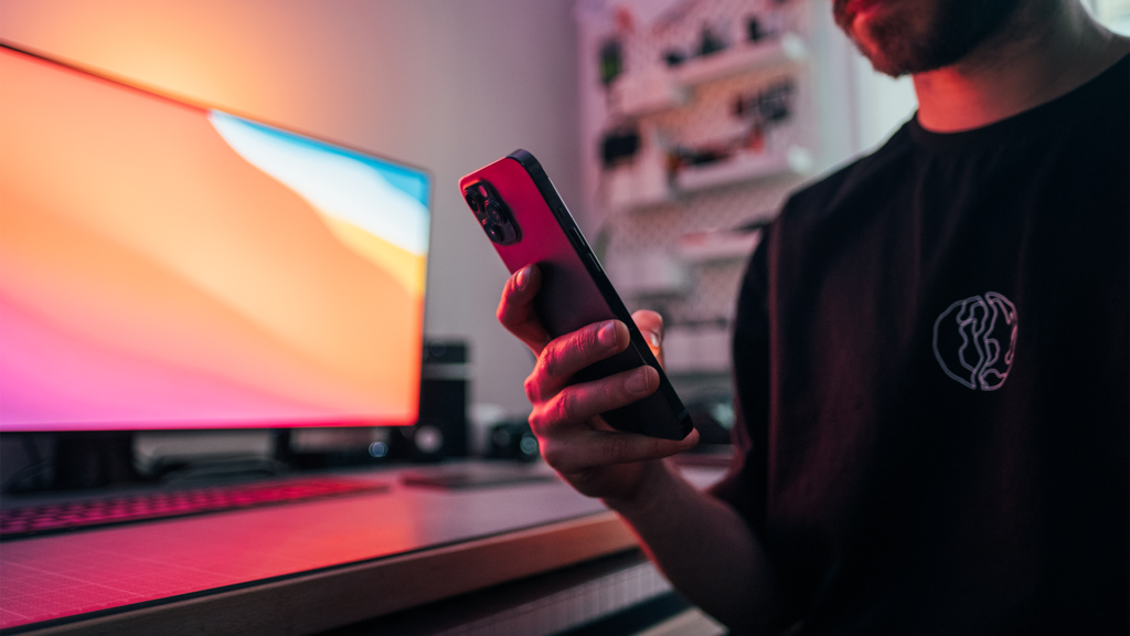 Mann wird von Computer-Bildschirm angeleuchtet und schaut dabei auf sein Smartphone.