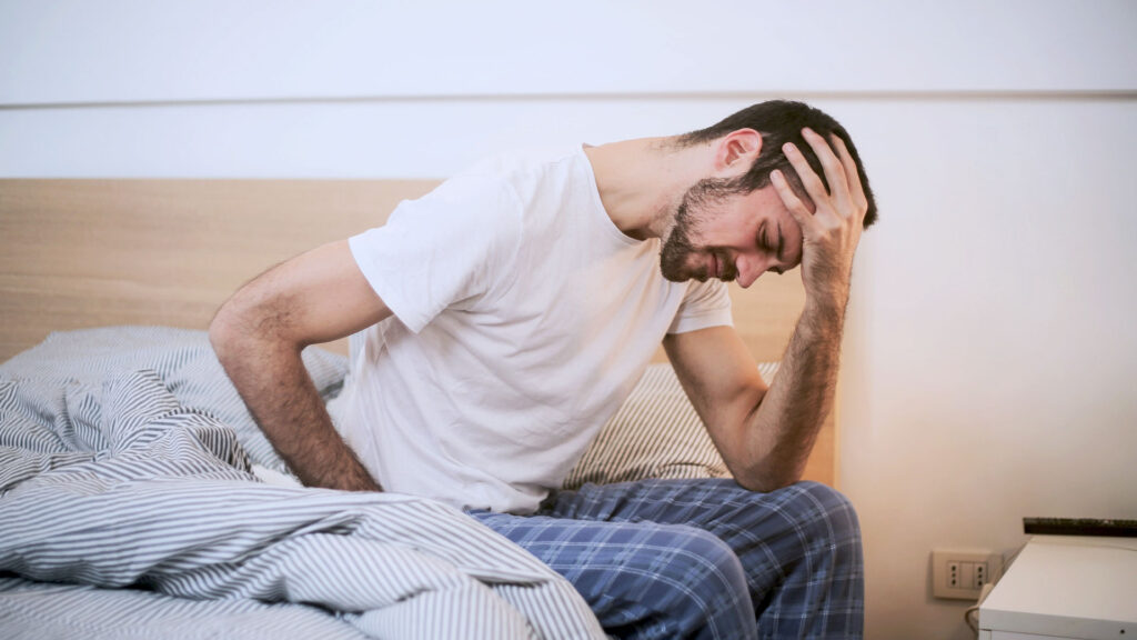 Mann sitzt auf dem Bett und hält sich die Stirn vor Schmerzen
