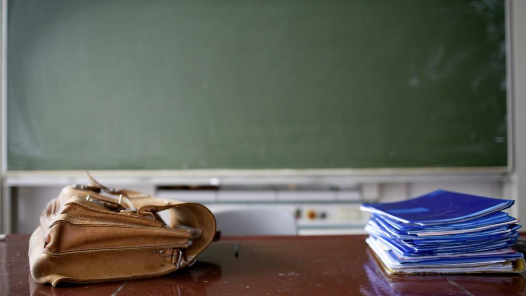 Klassenraum mit Lehrerpult auf dem blaue Arbeitshefte und eine braune Aktentasche liegen. Im Hintergrund eine grüne Tafel.