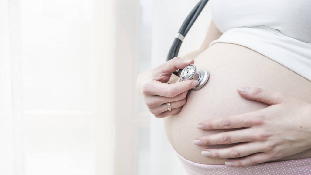 Schwangere hört ihren eigenen Bauch mit einem Stetoskop ab. Der Hintergrund ist hell und leicht verschwommen.