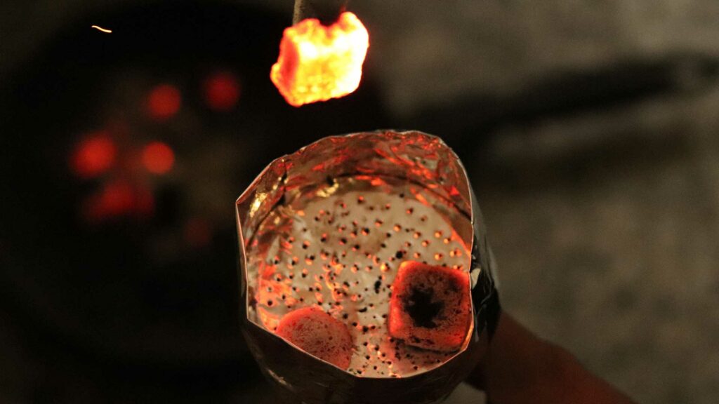 Ein glühendes Stück Shisha-Kohle wird gerade mit einer Zange aus einer Schale genommen.