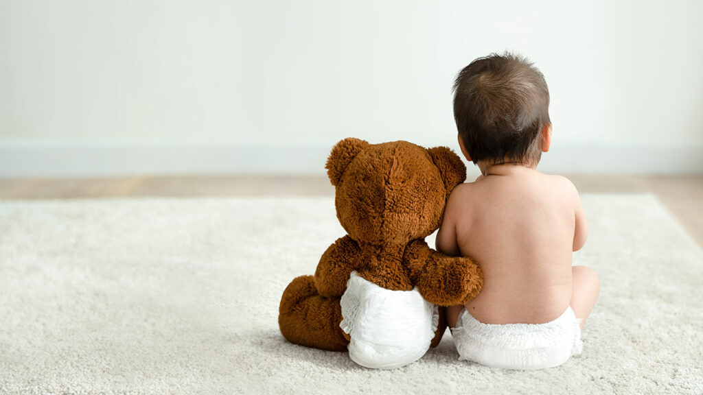 Ein Kleinkind und ein Teddybär in Windeln sitzen mit dem Rücken zur Kamera.