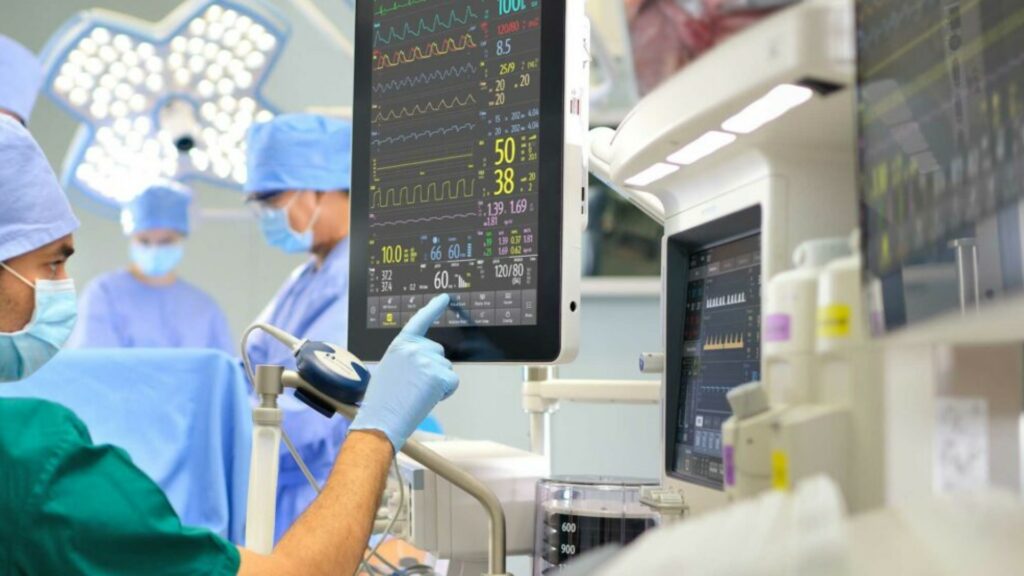 Ein Arzt überwacht im Operationssaal die Werter der operierten Person. Im Hintergrund sind Chirurgen während der Operation zu sehen.
