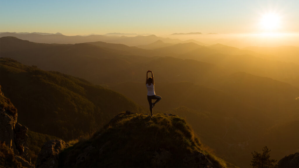 Frau macht auf einem Berg Yoga, die Sonne geht vor ihr unter.
