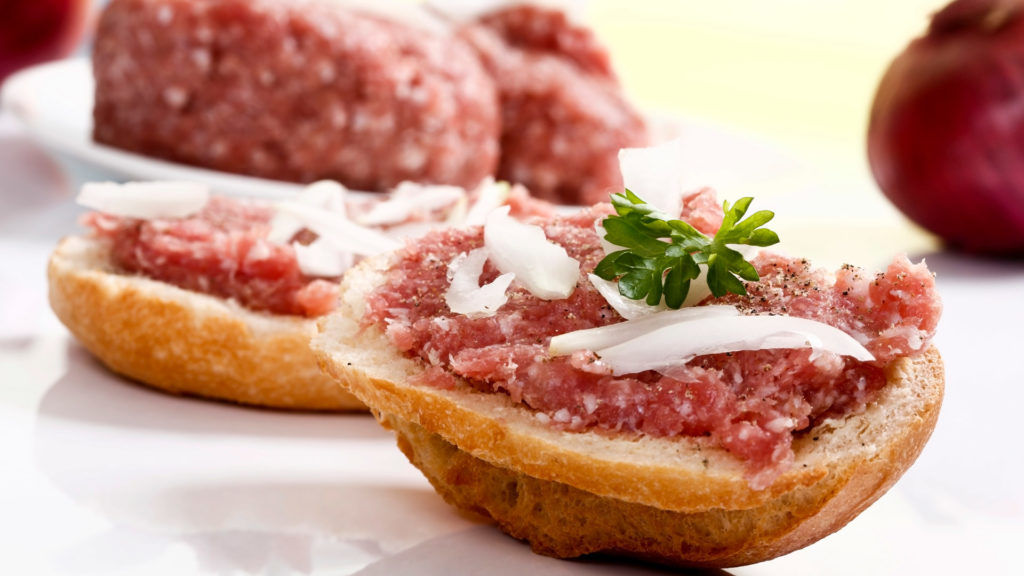Mett auf Brötchenhälften mit Zwiebelstücken und Petersilie darauf. Im Hintergrund ein Haufen Mett und eine ganze rote Zwiebel.
