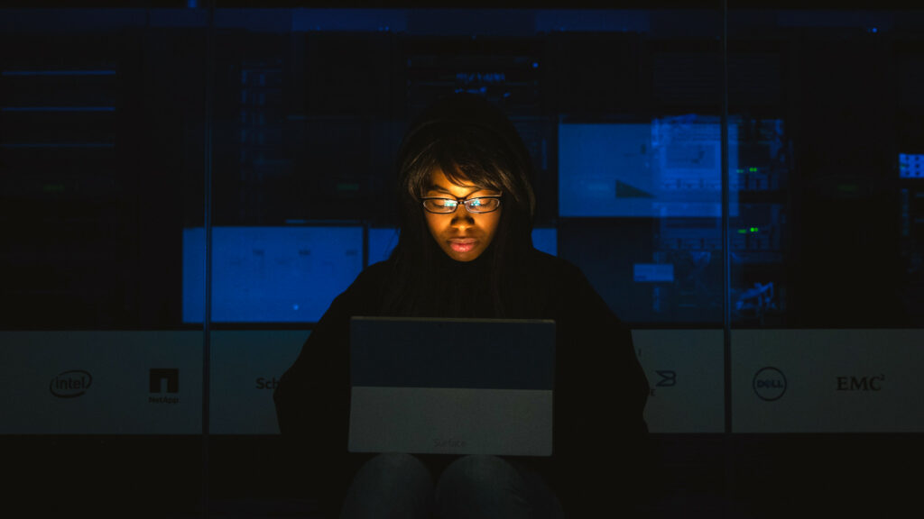 Eine Frau mit Brille sitzt in einer dunklen Umgebung mit einem Laptop auf dem Schoß. Das Licht des Bildschirms erhellt ihr Gesicht, während der Hintergrund kühl und dunkel bleibt.