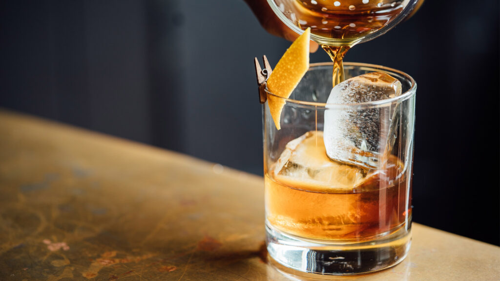 Whiskey wird in ein Glas eingeschenkt. Das Glas ist mit ein Orangenschale dekoriert worden und enthält Eiswürfel.