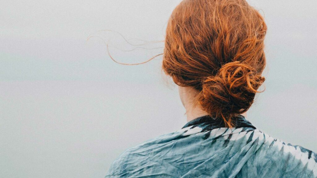 Zu sehen ist eine Frau mit rooten Haaren, die ihre Haare zum Dutt trägt. Sie kehrt der Kamera den Rücken zu, ihre Haaren wehen im Wind