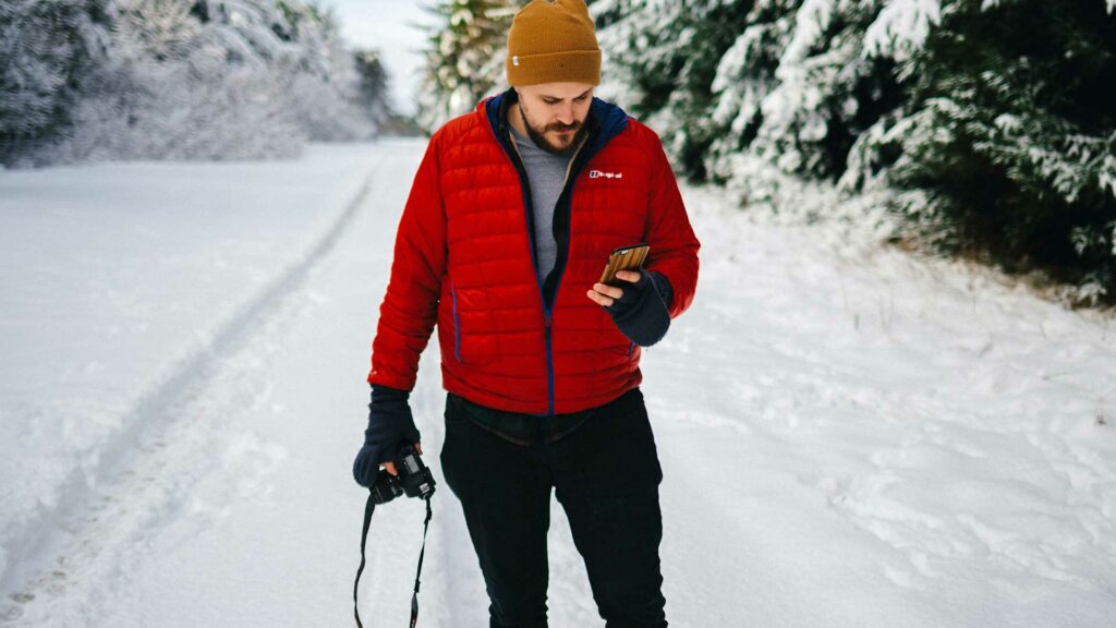 Ein Mann steht auf einem verschneiten Gehweg, umgeben von vielen Bäumen, und schaut auf sein Handy.