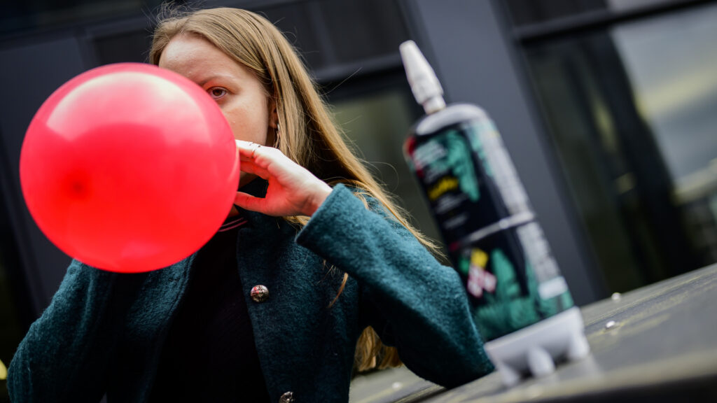 Eine Frau inhaliert den Inhalt eines Luftballons. Neben ihr steht eine Flasche Lachgas.