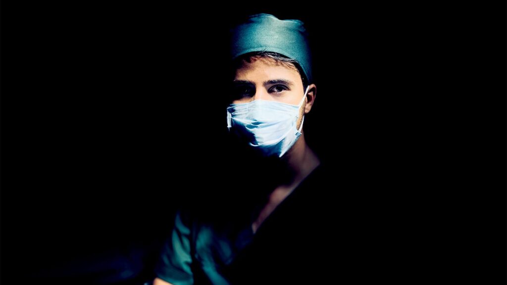 Junger Arzt mit Maske vor dunklem Hintergrund.