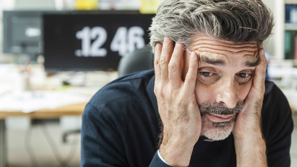 Mann mit grauen Haaren. Seine Stirn ist in Falten gelegt. Er hält erschöpft sein Gesicht.
