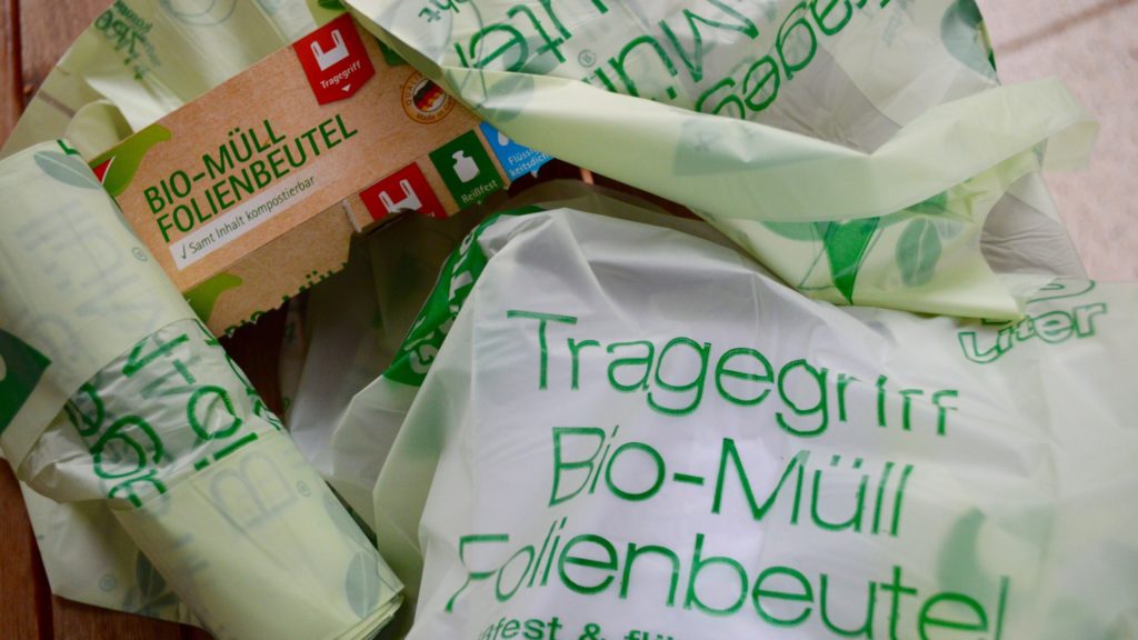 Eine Packung kompostierbare Bio-Müllbeutel aus Folie, umgeben von mehreren ausgebreiteten Beuteln mit grüner Aufschrift.