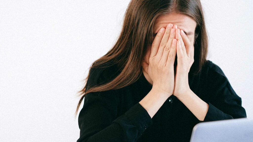 Frau sitzt erschöpft am Schreibtisch und hält sich die Hände vor das Gesicht. Ihre Stirn bildet, durch die dagegengedrückten Finger, Falten.
