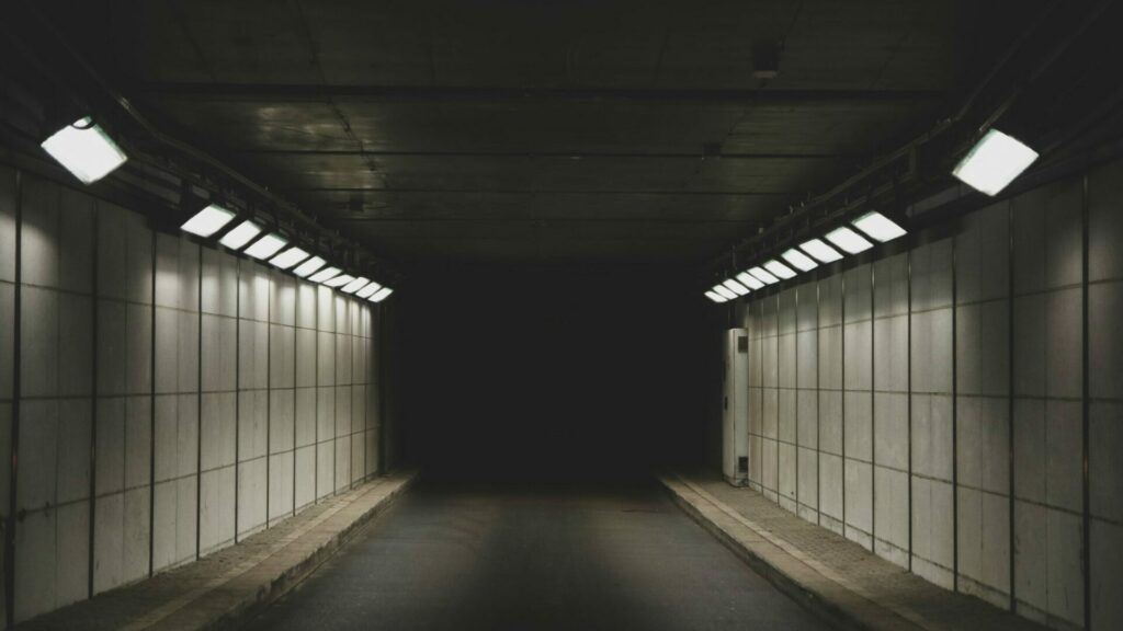 Ein Tunnel. Am Ende des Tunnels Dunkelheit. An den Seiten sind Lichtwerfer, beleuchten den Weg.