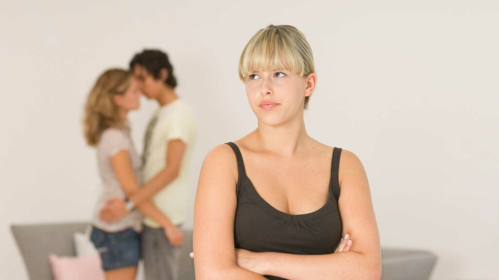 Eine Jugendliche ist eifersüchtig auf ein verliebtes Pärchen. Bild: Mädchen, Arme verschränkt, traurig blickend, im Hintergrund Mann, Frau sich umarmend.