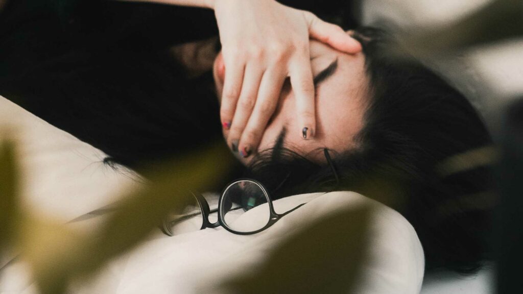 Eine Person liegt auf einem Kissen, bedeckt ihr Gesicht mit einer Hand und hat dunkle, leicht zerzauste Haare. Eine Brille liegt unscharf im Vordergrund.