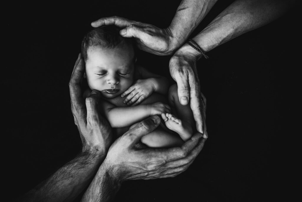 Neugeborenes Baby schläft, von mehreren Händen in einer Schwarz-Weiß-Aufnahme gehalten.