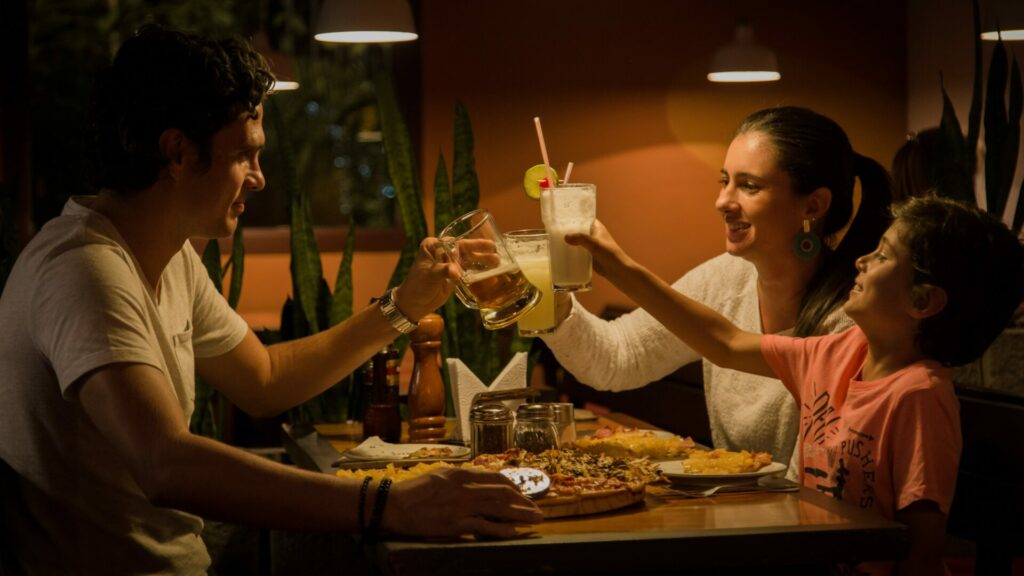 Eine Familie sitzt an einem Tisch in einem Restaurant und stößt mit ihren Getränken an. Sie lachen und scheinen eine Mahlzeit zu genießen. Der Tisch ist mit Pizza und anderen Speisen gedeckt, während warme Beleuchtung eine gemütliche Atmosphäre schafft.
