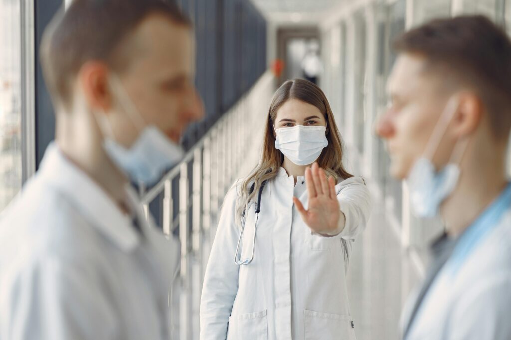 Eine Frau in einem weißen Kittel und mit Atemschutzmaske signalisiert zwei Männern: Halt!
