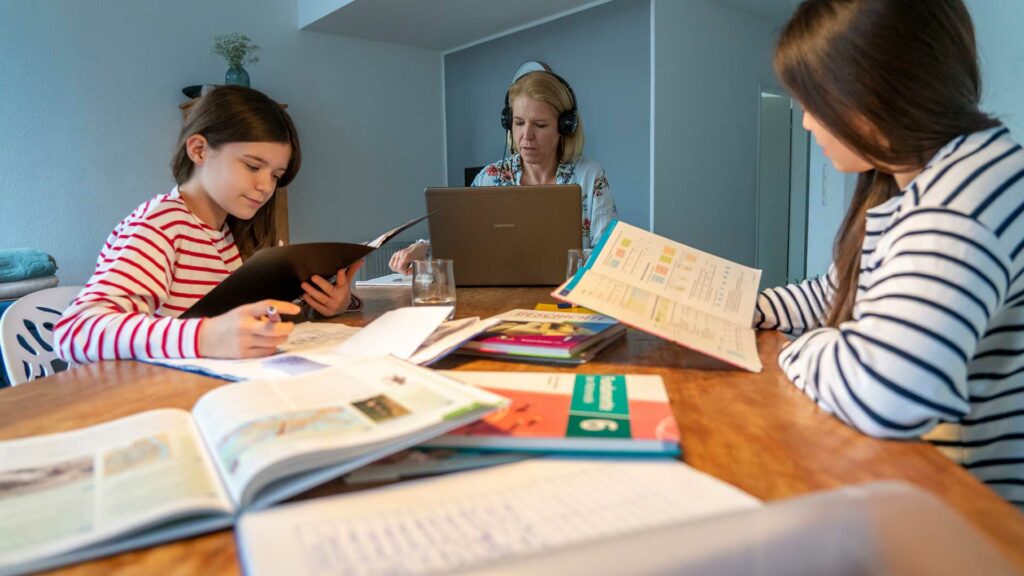 Zwei Kinder lernen Zuhause am Essenstisch für die Schule, ihre Mutter ist im Homeoffice und arbeitet am Laptop mit Videochat.