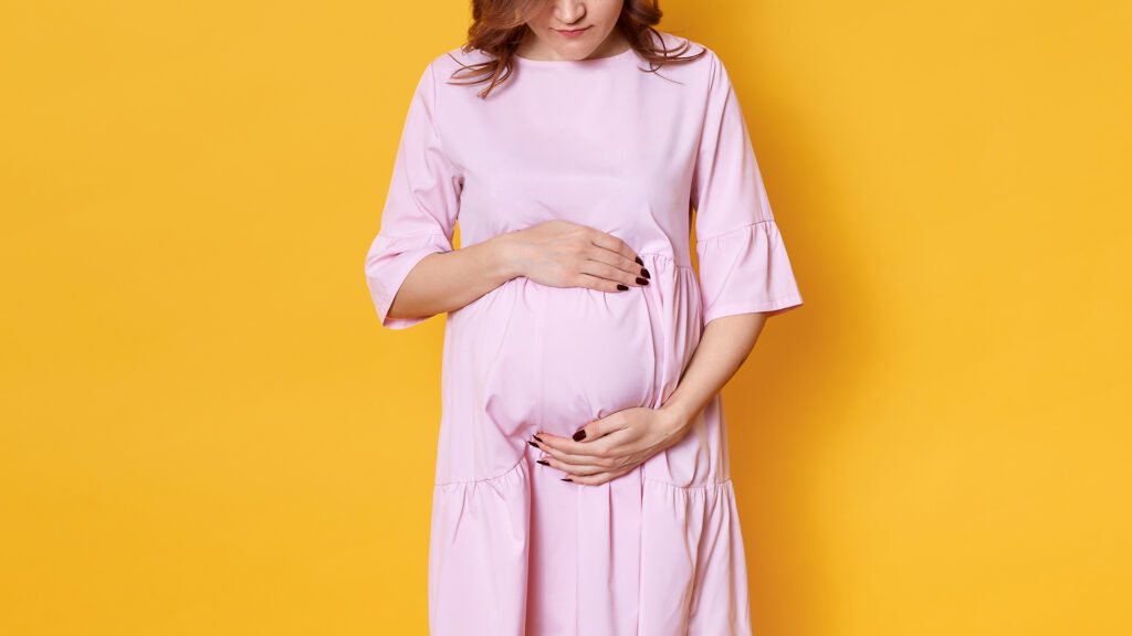 Eine hochschwangere Frau vor einer gelben Wand. Sie trägt ein rosafarbenes Kleid und umgreift ihren Bauch.