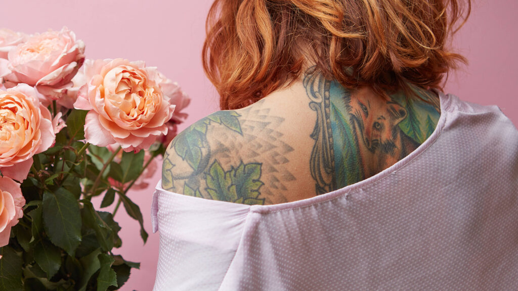 Frau mit tätowierter Schulter hält rosa Rosenstrauß vor rosa Hintergrund.
